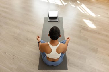 Meditasyon yapan yoga pozu veren bir kadın laptopun önünde minderin üzerinde oturur, muhtemelen sakin bir ev ortamında, güneş ışığının içinden geçtiği internet rehberli bir oturumu takip eder.