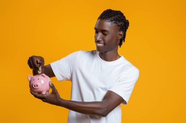 Akıllı Tasarruf. Piggy Bank 'a madeni para sokan, sarı stüdyo geçmişine poz veren, ekonomiden, planlama bütçesinden ve yatırımlardan keyif alan Afrikalı Amerikalı genç adamın portresi.