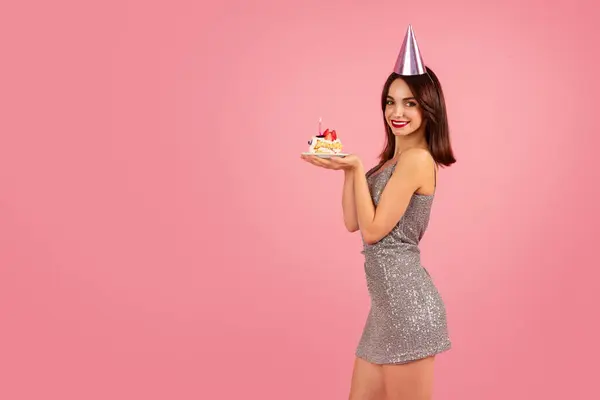 光沢のある銀製のパーティードレスとマッチングする円錐帽子を着たブラウンヘアの輝く女性は ピンクの背景にライトキャンドル付きのバースデーケーキを保持しています ホリデーイベント — ストック写真