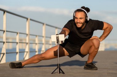 Aktif giyimli sporcu, deniz kenarındaki aerobikle akıllı telefonundaki egzersiz videosunu birleştiriyor ve sağlıklı vücut ve yaşam tarzını korumak için mobil teknolojinin kolaylığını vurguluyor.