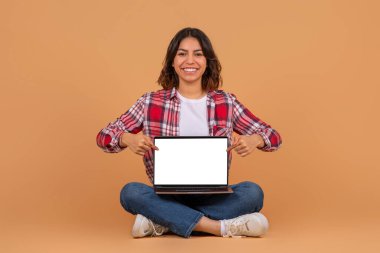 Sıradan giyinmiş mutlu bir orta doğulu kadın, boş ekranlı dizüstü bilgisayarı işaret ederek, renkli arka planda izole bir şekilde yerde oturuyor. Blogcu öneri ve çalışma, çalışma, harika reklam ve teklif
