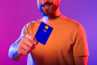 Milenyum adamı mor neon arka planda mavi plastik kredi kartı gösteriyor. Kesilmiş atış. Sanal bankacılık, sınırsız ödeme seçenekleri ve çevrimiçi finans hizmetleri reklamcısı.