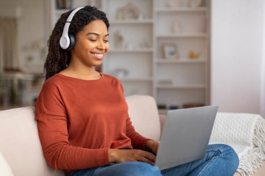 Gülümseyen siyah bir kadın kulaklıkla müzik dinlerken evde dizüstü bilgisayar başında mutlu bir Afrikalı Amerikalı serbest yazar kadın rahat bir şekilde oturma odasında kanepede oturuyor.