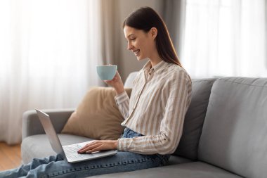 Genç serbest çalışan bir kadın dizüstü bilgisayarla çalışırken kahve içip eğleniyor. Parlak, havadar oturma odasında gri koltukta oturuyor.