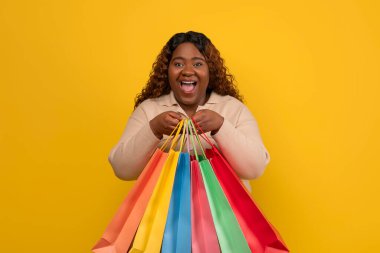 Milenyum aşırı kilolu Afrikalı Amerikalı kadın elinde renkli kağıt torbalarla alışverişin keyfini çıkarıyor. Stüdyo sarı arka planda izole edilmiş.