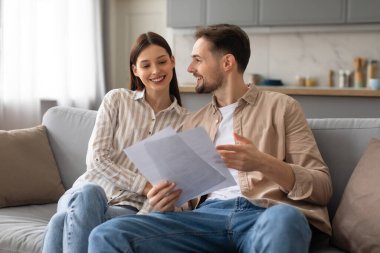 Kanepede yan yana oturan gülümseyen genç eşler, kağıtlar üzerinde olumlu bir tartışmaya giriştiler, evdeki rahat oturma odasında planlama ya da anlaşmaya işaret ediyorlar.