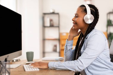 Saç örgülü genç kız kulaklık kullanıyor, dikkatle boş bilgisayar monitörüne bakıyor, evdeki masasından online bir çalışma seansına katılıyor.
