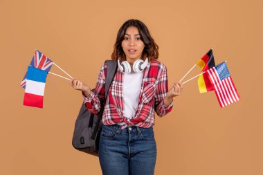 Kafası karışmış orta doğulu bir kadın, kulaklıkları ve sırt çantalı bir öğrenci elinde farklı ülkelerin bayraklarıyla kameralara bakıyor, bej arka plan, yurtdışında eğitim için ülke seçiyor.