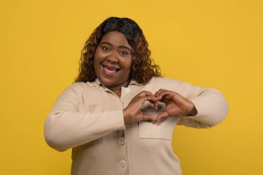 Sevimli, tombul, güzel, bin yıllık Afro-Amerikan kadın bej gömlek giyiyor, sevgi jestini kalbe gösteriyor ve kameraya gülümsüyor, sarı stüdyo arka planında izole bir şekilde poz veriyor.