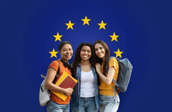 Three joyful millennial students with backpacks and books hugging and smiling against a blue background with the European Union stars, symbolizing friendship and education