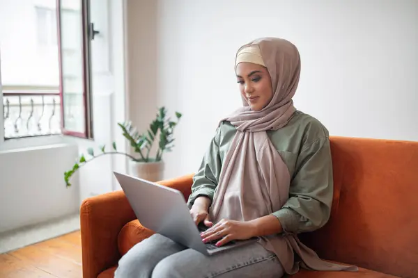 Work and Learn. Islamic millennial woman in headscarf works on her laptop, combining remote career and e-learning, sitting on sofa with computer in cozy modern living room at home