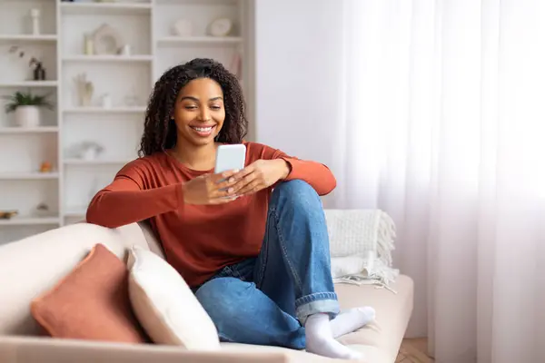 Black smiling woman in casual wear using her smartphone at home, happy african american female comfortably sitting on couch in living room, browsing new app on mobile phone, free space