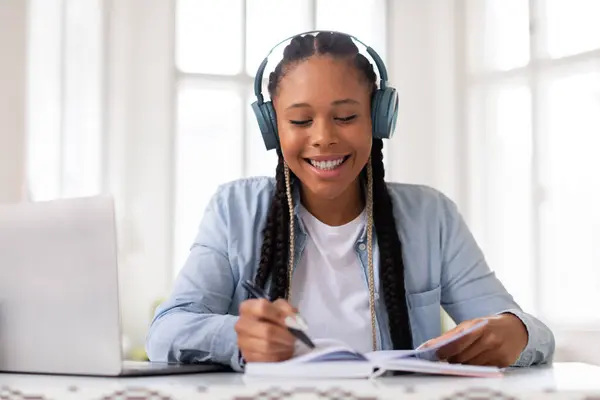 微笑着的黑人少女喜欢带着耳机学习 在明亮 阳光灿烂的房间里从笔记本电脑上记笔记 体现了富有成效的远程教育 — 图库照片