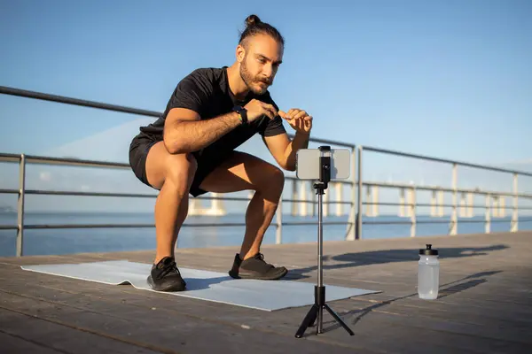 暑期训练 身穿运动服的运动员博主在海边蹲着 在他的智能手机上流着锻炼视频以获得动力 将户外运动与技术结合起来 Fitness博客 — 图库照片