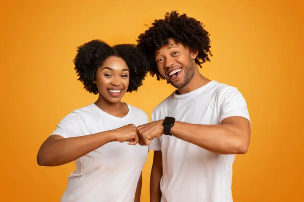 Cheerful young african american man and woman friends or lovers wearing white t-shirts doing fist bump and smiling at camera, celebrating success, showing teamwork, orange background