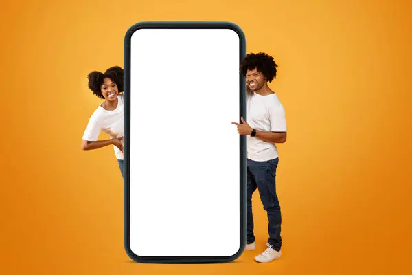 Nice online offer. Cool stylish young african american man and woman pointing at big smartphone with white blank screen mockup copy space between them, orange background, web-banner