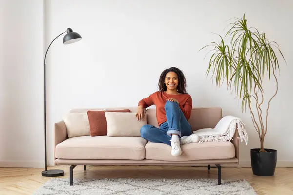 Relaxed young black woman with joyful smile sitting comfortably on beige sofa at home, happy millennial african american lady posing in well-lit living room, exuding casual elegance, copy space