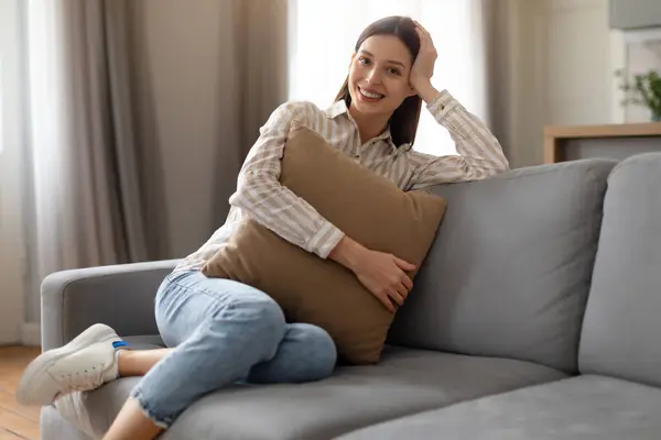 穿着条纹衬衫的快乐女人 抱着心满意足的垫子 在明亮 通风的客厅里盘腿坐在柔软的沙发上 — 图库照片