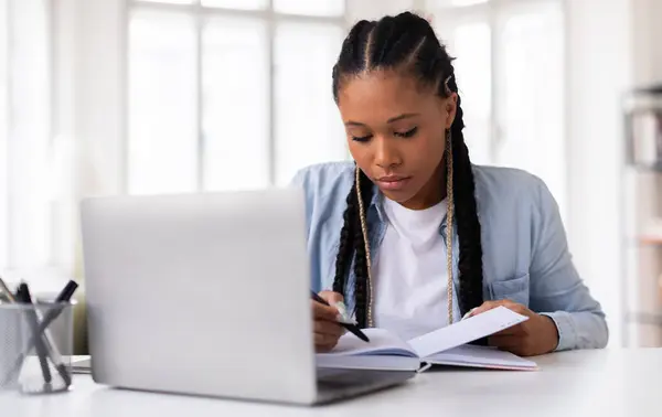 Skoncentrowany Czarny Student Warkoczami Studiuje Uważnie Pisząc Notatniku Przy Biurku — Zdjęcie stockowe