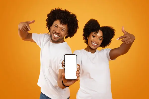 Cool cheerful millennial black couple man and woman wearing white t-shirts holding smartphone with white blank screen mockup copy space for mobile app or online offer advertisement