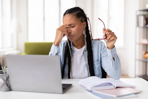頭痛に悩まされた黒人女性の学生は 机の上のラップトップで勉強しながら 鼻の橋をマッサージし 眼鏡を保持しながら ホームインテリア — ストック写真