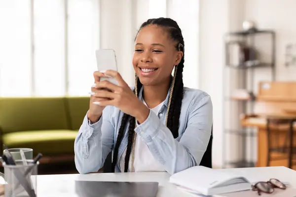 Engaged and happy black teen lady with braided hair using her smartphone, possibly taking break from studying or communicating with friends
