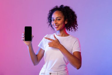 Kıvırcık saçlı neşeli siyahi kadın boş akıllı telefon ekranını işaret ediyor. Canlı pembe ve mavi ışıklı arka plana karşı yeni bir uygulama sunuyor.