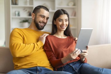 Harika bir uygulama. Mutlu Genç Çift Evde Dijital Tablet Kullanıyor, Gülümseyen Milenyum Eşleri Oturma Odasında Modern Gadget ile Dinleniyor, İnternete ya da İnternetten Alışveriş Yapıyor