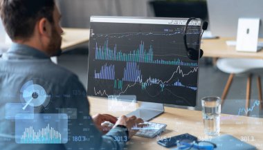 Odaklanmış bir adam, bir ofis ortamında bir kulaklıkla donatılmış bir bilgisayar monitörü üzerindeki karmaşık mali istatistikleri analiz ediyor.
