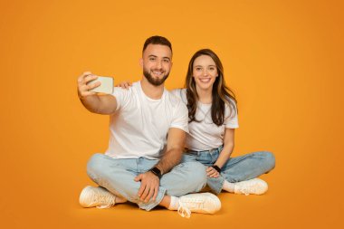 Beyaz tişört ve mavi kot pantolon giymiş, yerde bağdaş kurup, akıllı telefonlarıyla selfie çeken beyaz tenli ve turuncu renkli bir çift.