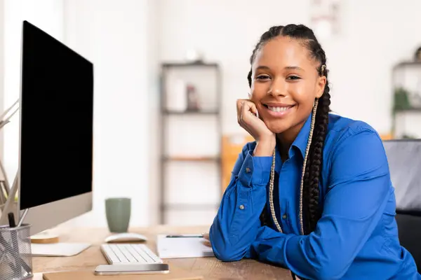 青いシャツの陽気な黒人女性学生は きれいなコンピュータデスクで勉強セッションを楽しんで 空白のコンピュータ画面とリラックスしたポーズでカメラで笑顔 — ストック写真