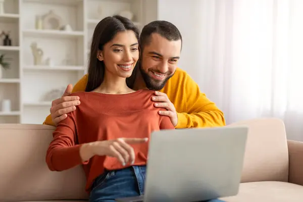 Happy Young Spouses Using Laptop At Home, Booking Vacation Together, Smiling Beautiful Man And Woman Planning Trip, Making Hotel Reservation Online, Purchasing Flight Tickets, Husband Embracing Wife