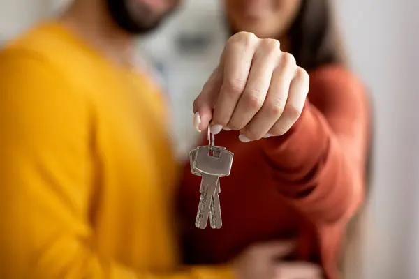 Closeup Shot Of Romantic Young Couple Holding Home Keys In Hands, Loving Millennial Spouses Celebrating Moving To Their Own House, Embracing And Smiling, Cropped Image With Selective Focus