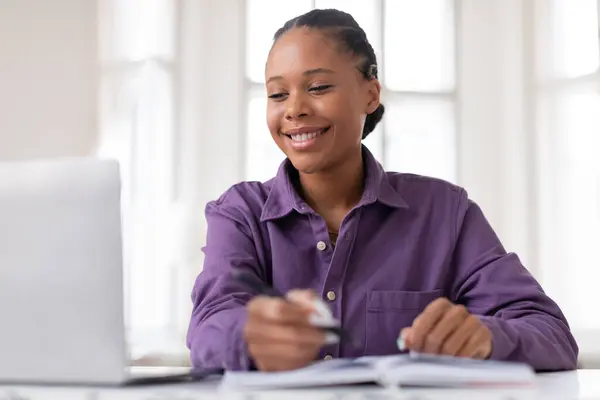 穿着紫色衬衫 心满意足的黑人女生 一边在笔记本电脑上记笔记 一边带着灿烂的笑容 坐在家里光线充足的房间里学习 图库照片