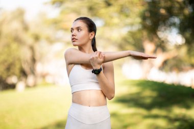 Huzurlu bir parkta, şık spor kıyafetleri içinde kendini adamış beyaz bir kadın, şehir parkının dışında, teknoloji meraklısı bir fitness rutinini ima ederek kol esneme hareketi yapıyor.
