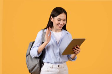 Meşgul bayan öğrenci dalgaları merhaba derken tabletine odaklanıyor, arkadaşlığı teknolojiyle birleştiriyor sıcak sarı arka plana karşı, e-öğrenmeyi sembolize ediyor