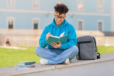 Neşeli genç Brezilyalı öğrenci öğrenim notları alıyor, çalışma kitapları ve sırt çantalarıyla dışarıda, üniversite parkında ders çalışıyor, eğitim keyfi yapıyor.