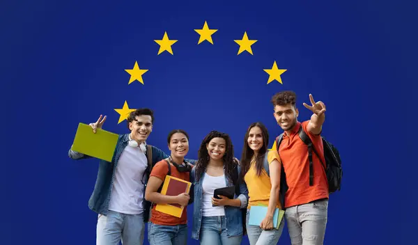 Group of five young and diverse students happily showing victory signs and holding study materials against a blue background with the European Union stars, studio, panorama