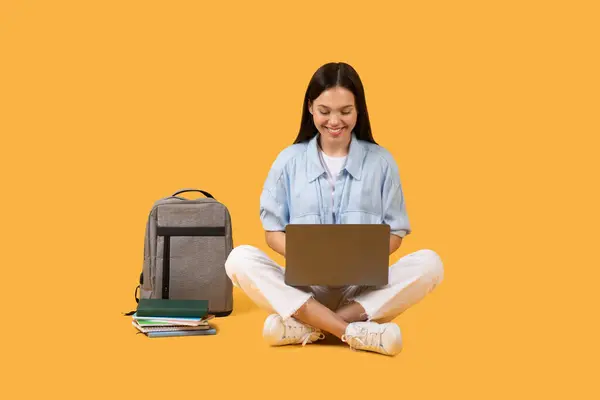 カジュアルな服装の女性学生は 彼女の膝の上にラップトップと近くのバックパックで横足し 黄色の背景に対して穏やかな研究環境を表示 — ストック写真