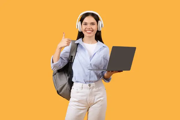 快乐的女学生拿着笔记本电脑 戴着耳机时放弃了大拇指 象征着在一个黄色背景下的电子学习环境中的成功和认可 — 图库照片