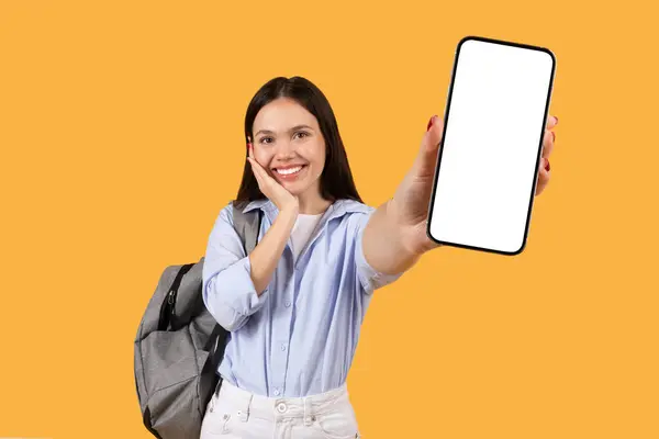 白いスマートフォンのスクリーンを示し 黄色の背景に対して微笑んでいるバックパックが付いている幸せな女性の学生 教育用アプリやプラットフォームの広告に最適 — ストック写真