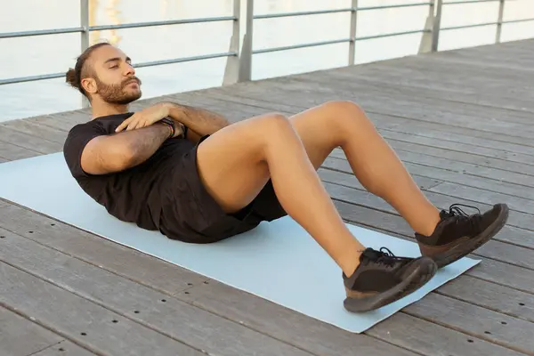 海滨健身 身穿运动服 充满热情的欧洲小伙子在自己的腹肌上做着仰卧起坐 在码头边进行户外活动 展示着健康而积极的生活方式 — 图库照片