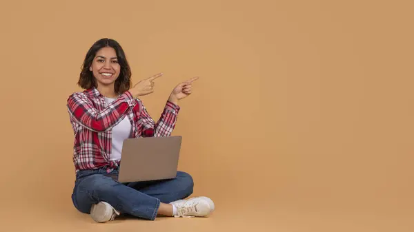 ベージュスタジオの背景の上に床に座ってラップトップコンピュータを持つ陽気な若い中東の女性学生 コピースペースを指し 素敵なオファーを示すパノラマ — ストック写真