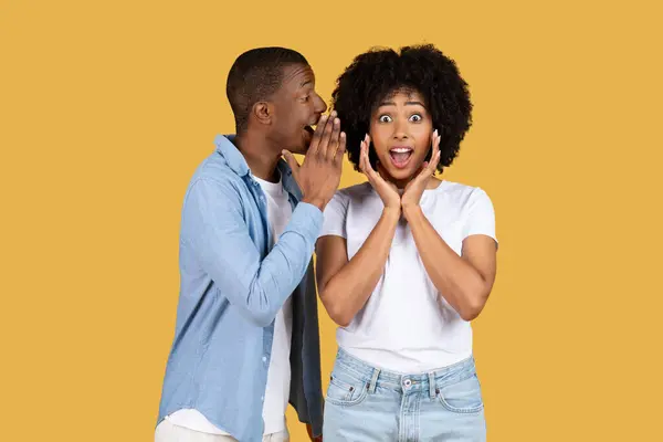 Excited millennial African American man whispering a secret into a womans ear, causing her to react with a surprised and joyful expression on a yellow background, studio