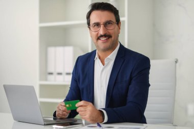 Koyu mavi takım elbiseli neşeli orta yaşlı Avrupalı işadamı, elinde parlak, modern ofis ortamında bir dizüstü bilgisayara online ödeme yapmaya hazır bir kredi kartı tutuyor. İş, iş finansmanı