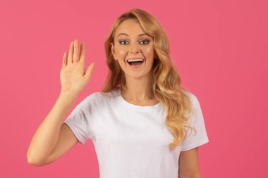 Dostça selamlar. Neşeli genç sarışın kadın pembe arka planda stüdyo portresiyle selam veriyor. Beyaz tişörtlü bir kadın selam veriyor, pozitif enerji yayıyor. İletişim konsepti