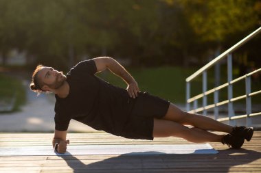 Açık hava antrenmanı. Spor kıyafetli kaslı adam yan dirsek tahtası egzersizi yapıyor, dışarıda minder üzerinde çalışıyor, uzun bir atış. Çekirdek kaslarını güçlendirmek için antrenman, spor motivasyonu