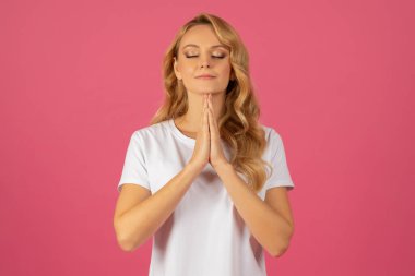 Sarışın genç bayan dua eden ellerini sıkıca tutarak dua ediyor. Pembe stüdyoda gözleri kapalı, portre fotoğrafı çekiliyor. Umut kavramı, ruhani meditasyon ve inanç.