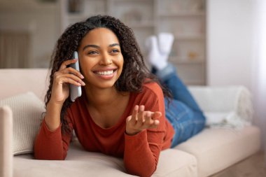 Evdeki koltukta uzanırken cep telefonuyla konuşan neşeli siyahi kadın, gülümseyen Afrikalı Amerikalı bir kadının Mutlu Cep Telefonu Konuşması, Oturma odasında dinlenme