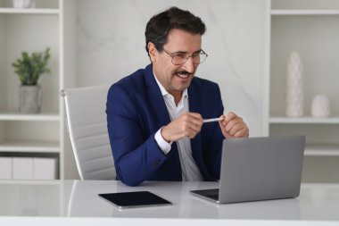 Mavi takım elbiseli komik Avrupalı orta yaşlı işadamı elinde kalem tutarken kahkahaya dalıyor, muhtemelen dijital bir konferansa tepki veriyor ya da bilgisayarındaki komik içeriğe gülüyor.